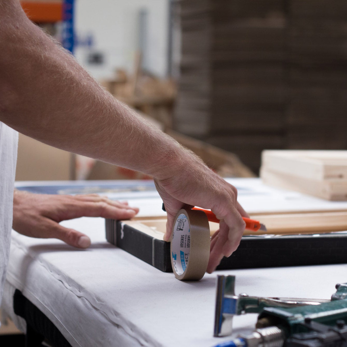 A close-up of a person's hands applying masking tape to a wooden frame on a workbench in a workshop setting.